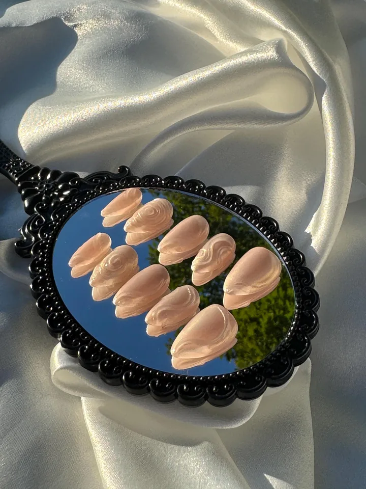 Ten pearly blush almond press on nails with raised flowing swirls, arranged on a black ornate mirror over ivory satin.