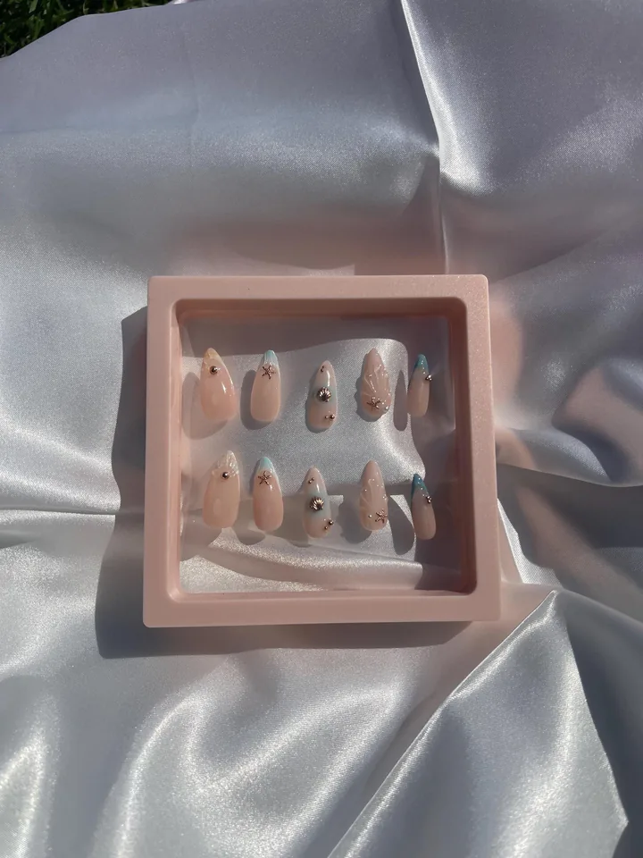Ten nude almond press on nails with pale blue accents, raised shell textures, tiny pearls and metallic shell and starfish charms in a pink display frame.