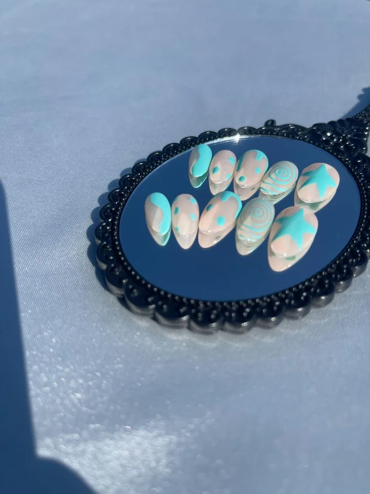 Ten nude almond press on nails with turquoise stars, spirals, dots and wavy shapes on a black ornate mirror.