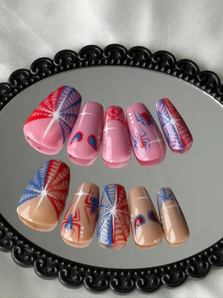Pink and nude coffin nails with red-and-blue spiderwebs, spider details and white starbursts on a black mirror